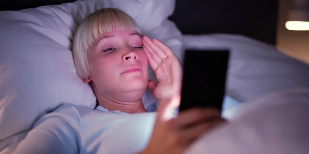 Woman in bed looking at her phone at night, blue light disrupting melatonin and sleep cycle.